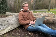 Calle Rosengren sitter utomhus med en dator och lutar sig mot en trädstam. Foto.