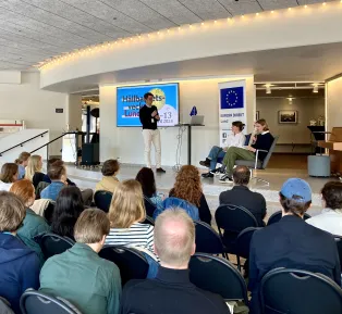 Tre personer talar från en scen inför en sittande publik på Botulfshörnan i Stadshallen. Foto.